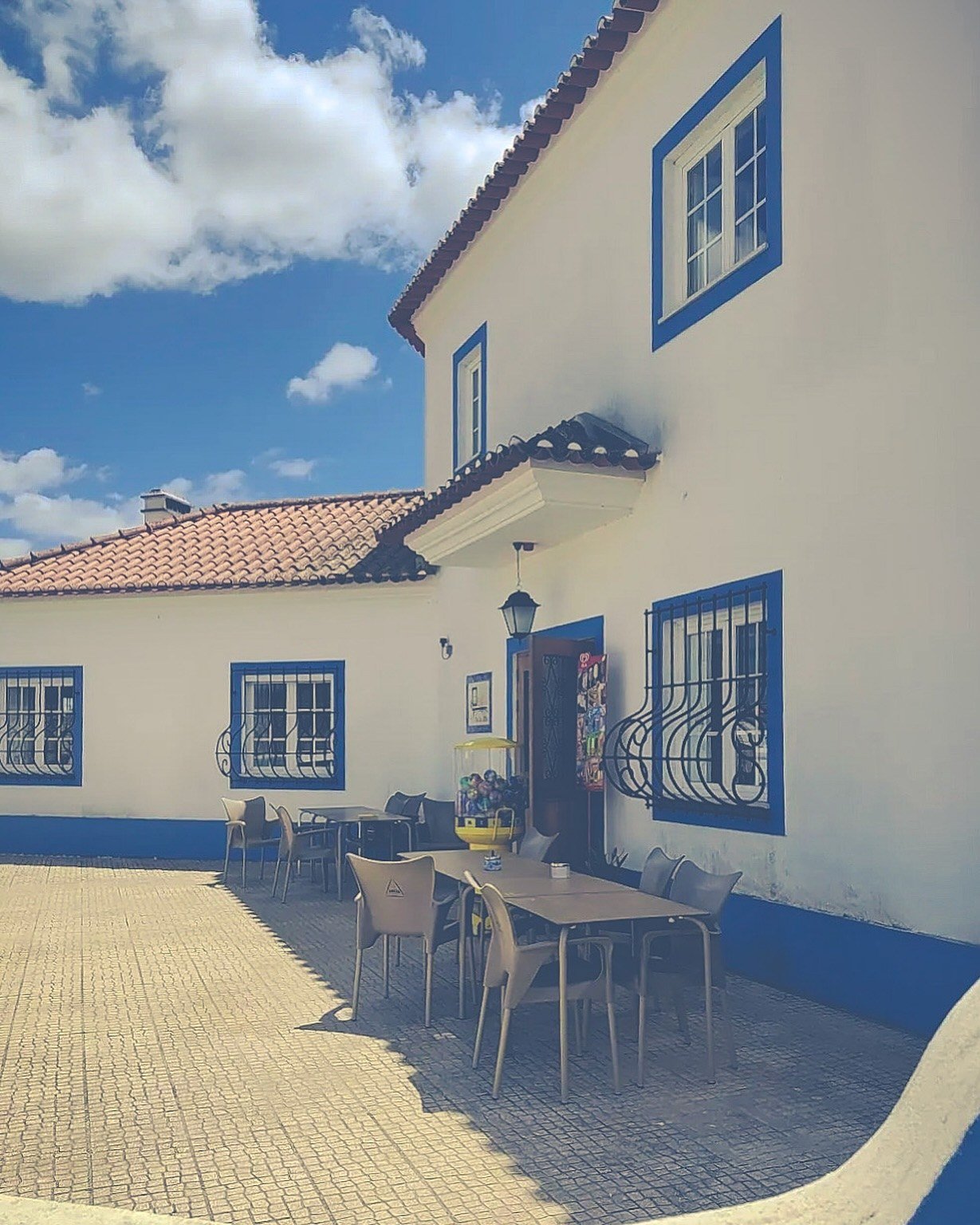 Fachada branca com barras azuis e mesas no pátio exterior do Pial das Bilhas